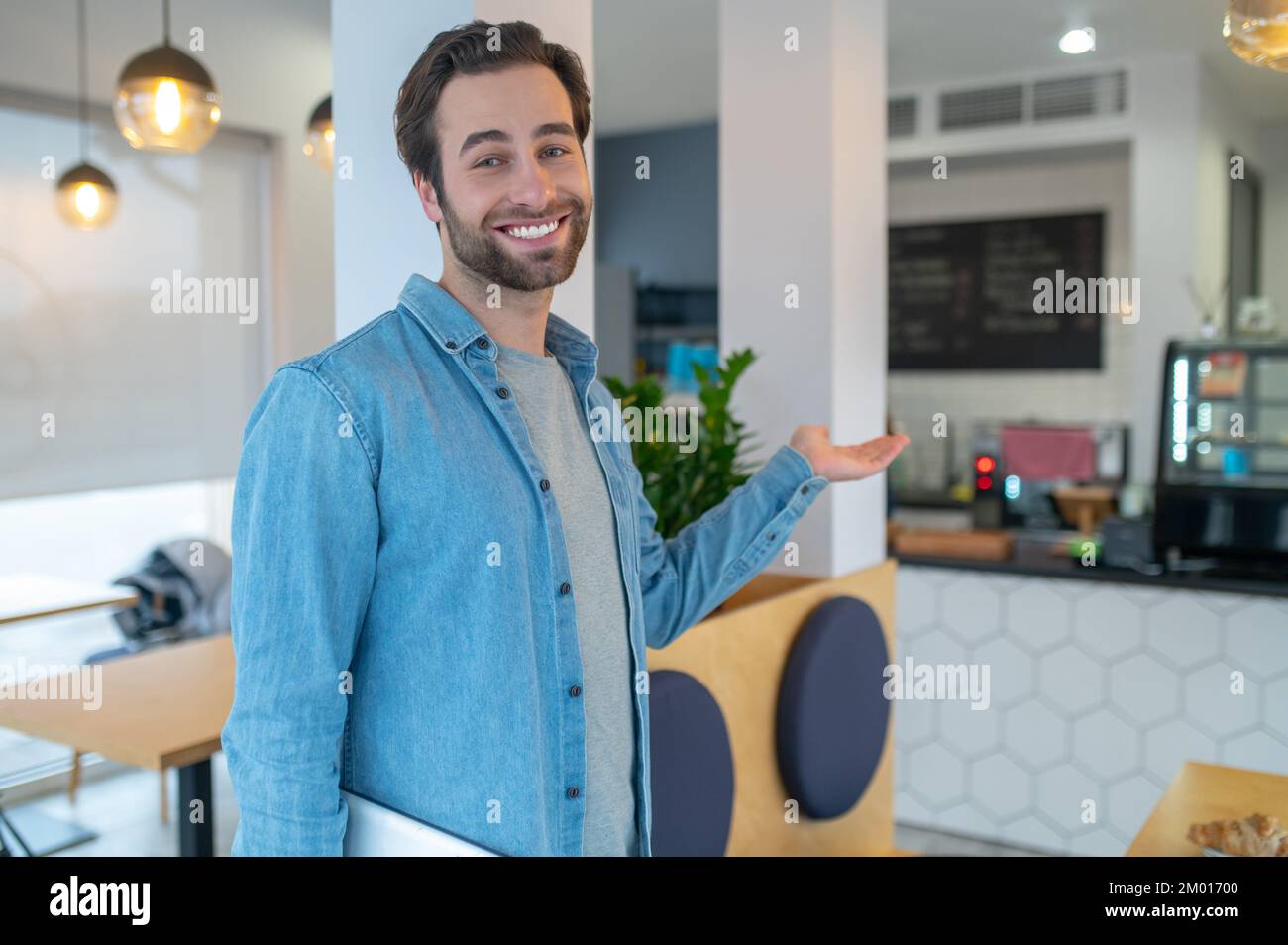 Invito. Attraente giovane uomo bearded con un sorriso toothy affably guardando la macchina fotografica con gesto invitante nel caffè. Foto Stock