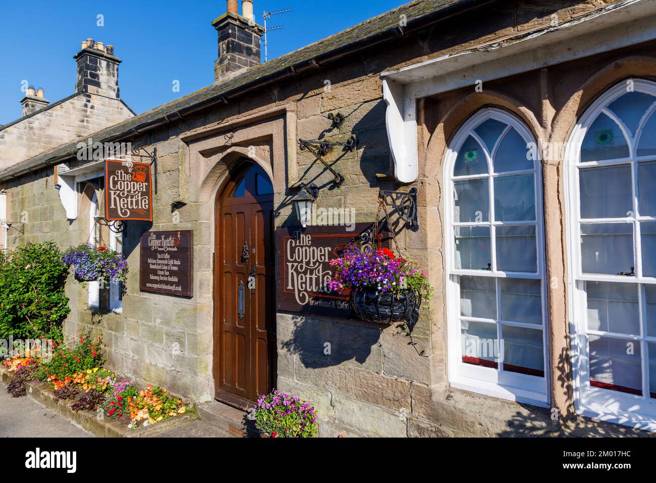 Il Copper Kettle, sale da tè tradizionali a Bamburgh, un villaggio del Northumberland sulla costa nord-orientale in una giornata di sole con fiori e cestini appesi Foto Stock