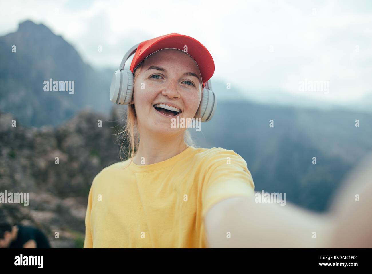 Giovane donna sorridente zaino in spalla con capelli corti brunette e beanie giallo, prendendo selfie con smartphone facendo segno di vittoria con mano in cima Foto Stock
