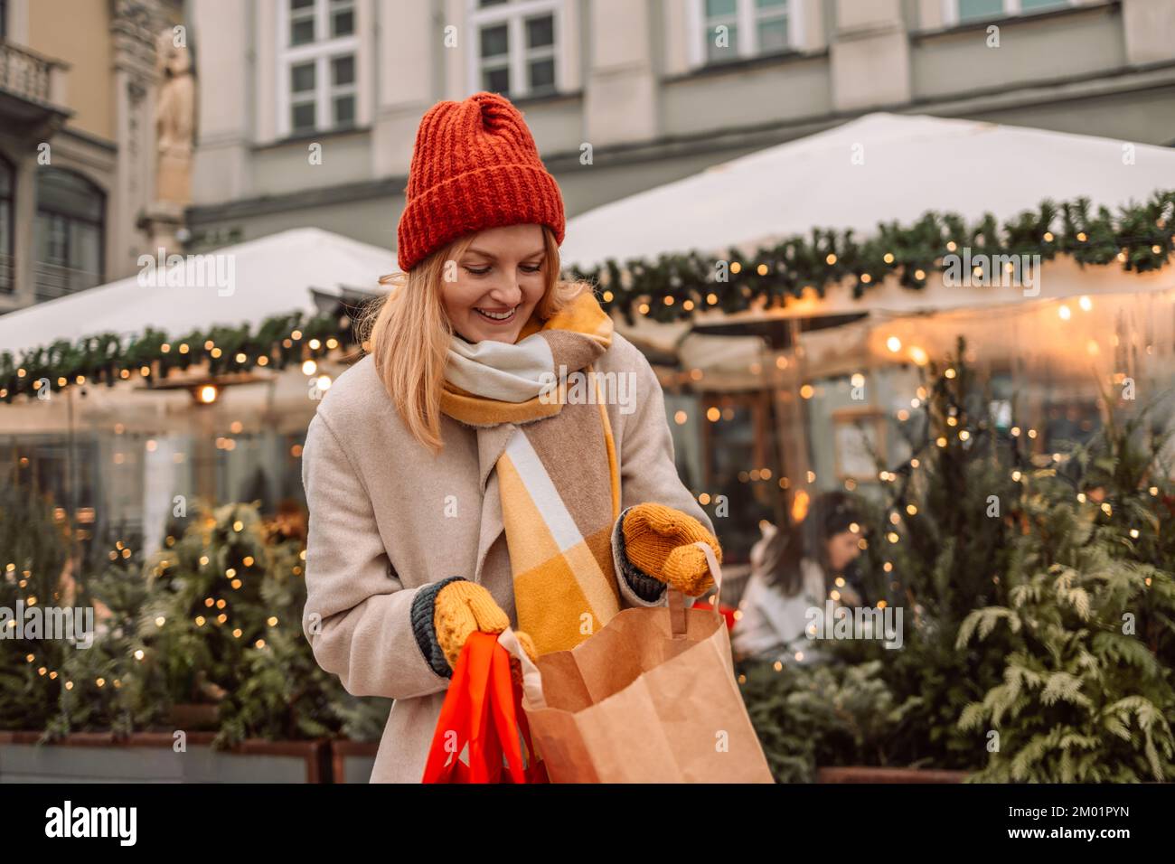 Attraente bionda giovane donna shopping, tenendo borse rosse shopping e scatole regalo con cappello rosso di Natale, sorridendo mentre si è in piedi di fronte al negozio Foto Stock