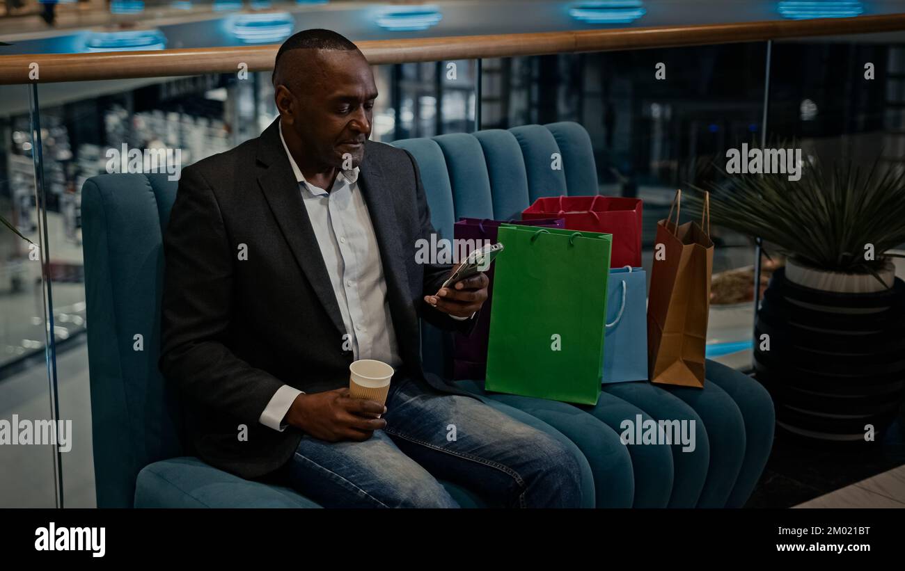 Uomo africano adulto uomo d'affari di mezza età con borse per la spesa nel negozio del centro commerciale con caffè tè tazza navigazione scorrimento telefono cellulare app di acquisto online Foto Stock