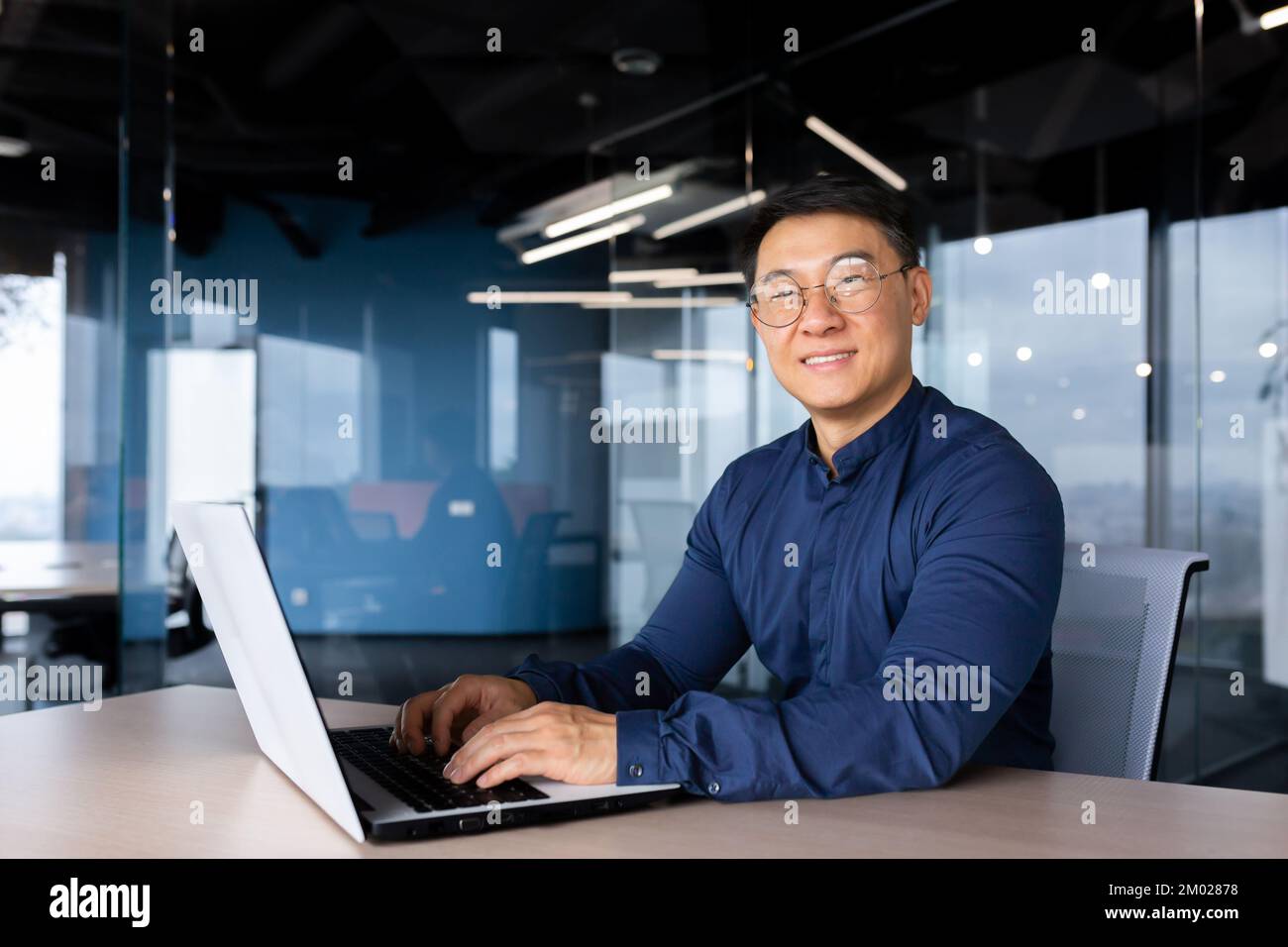 Uomo d'affari asiatico di successo sorridente e guardando la fotocamera, programmatore portiere all'interno dell'ufficio utilizzando il codice di scrittura del laptop per la sicurezza del software. Foto Stock