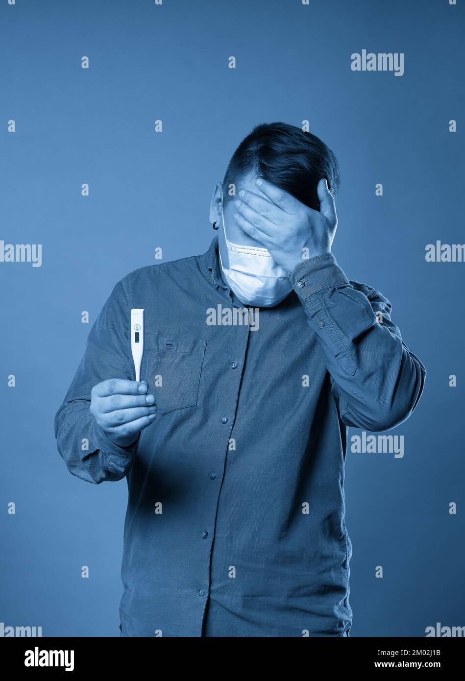 Giovane uomo in tessuto casual indossare maschera medica protettiva eccitato, stressato e spaventato dopo la lettura molto alta temperatura risultato da digitale Foto Stock