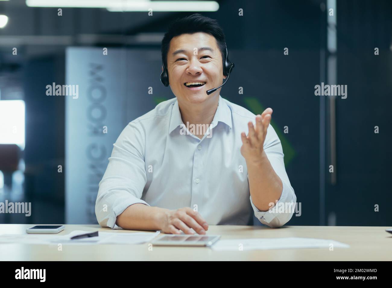 Call center. Telemarketing, assistenza clienti per la crescita delle vendite. Un giovane asiatico parla e consulta la fotocamera per il servizio clienti in cuffia. Seduto sorridendo in ufficio al tavolo. Foto Stock