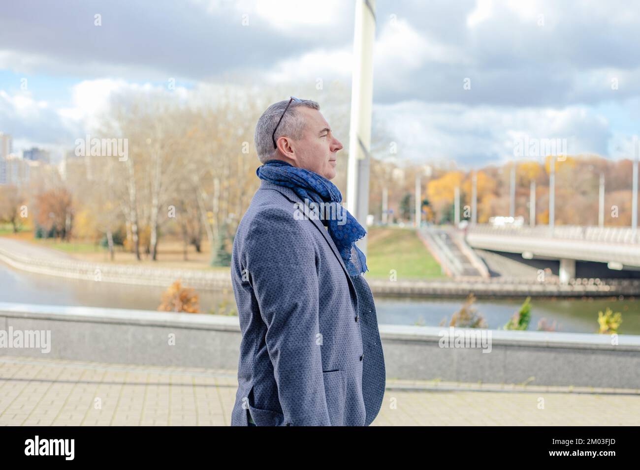 Vista laterale di un serio uomo di mezza età con capelli corti che indossa giacca grigia, sciarpa blu, in piedi su piastrelle vicino al fiume. Foto Stock