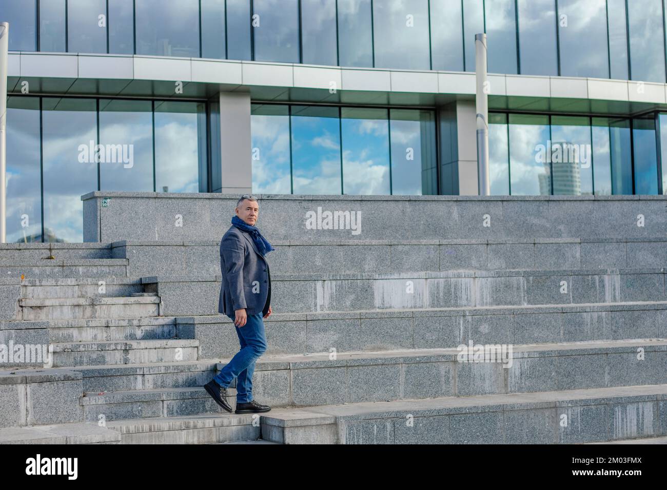 Vista laterale di un uomo di mezza età con capelli corti che scendono le scale in cemento dell'edificio moderno, guardando la macchina fotografica in città. Foto Stock