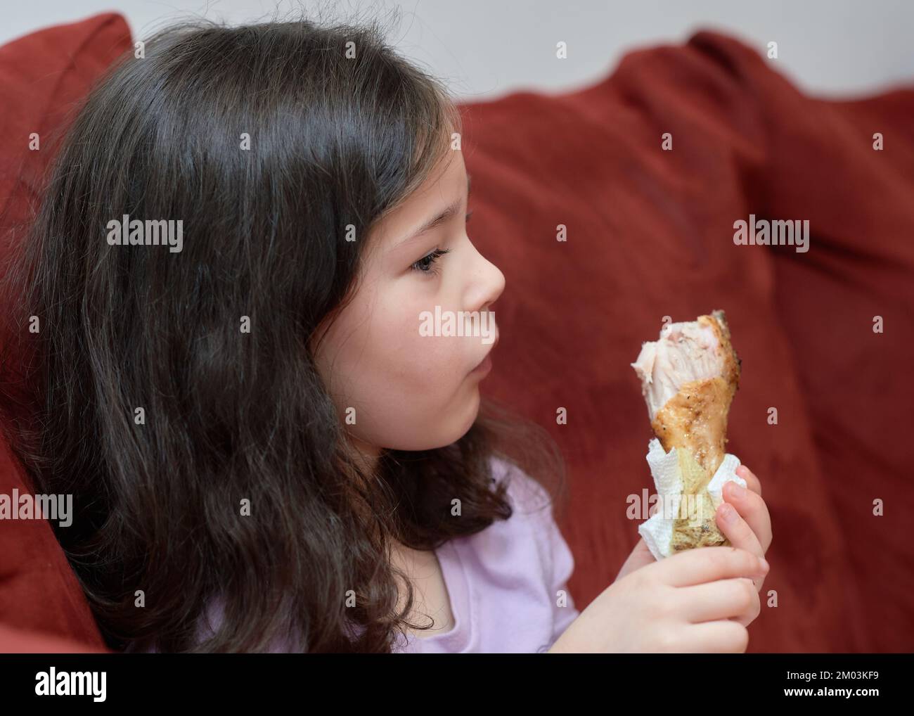 La giovane ragazza espressiva mangia pollo e verdure per cena sul divano mentre guarda la TV Foto Stock