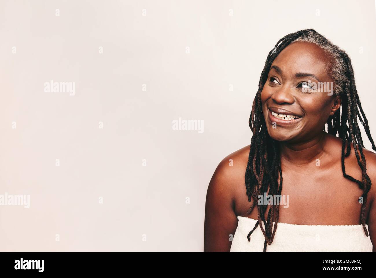 Donna rinfrescata che guarda via con un sorriso mentre avvolto in un asciugamano da bagno. Donna matura con Dreadlocks in piedi contro uno sfondo studio. Buon midd Foto Stock
