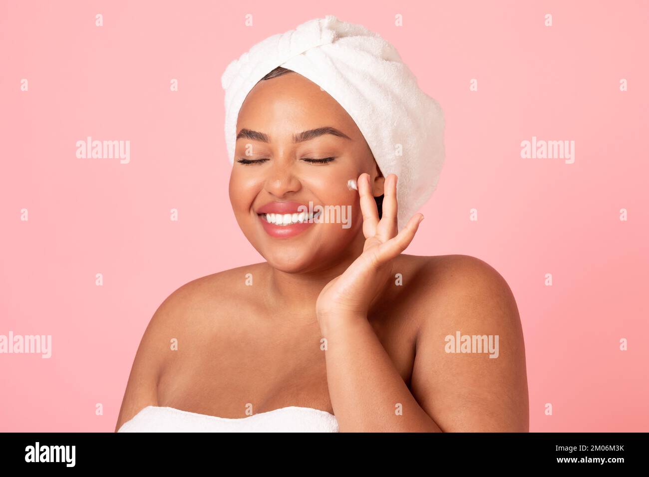 Felice corpo nero donna positiva applicare il prodotto di bellezza sul suo viso e sorridente, donna avvolto in asciugamano con crema viso Foto Stock