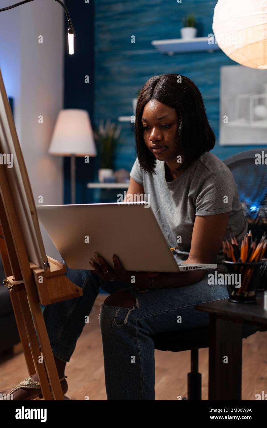Donna afroamericana con interessi artistici che usa il laptop per prendere lezioni d'arte tramite videoconferenza da studio creativo. Disegno studente cercando tecniche di schizzo su internet per fare grafica. Foto Stock
