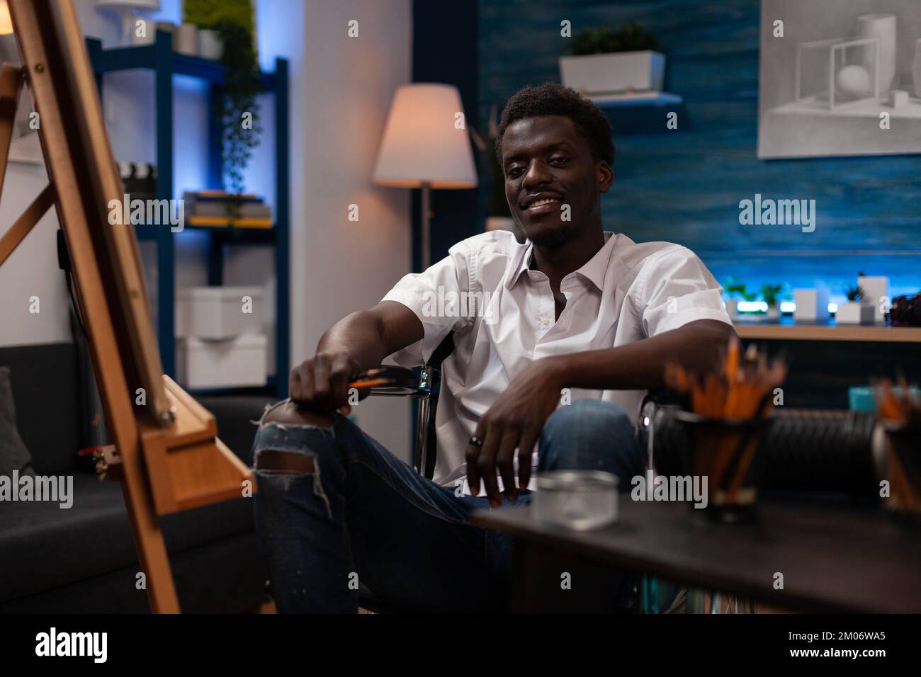 Artista sorridente con malattia degenerativa cronica in studio d'arte. Persona afroamericana con abilità artistiche vivere con motoneurone condizione medica tenendo matite colorate a casa atelier disegno. Foto Stock