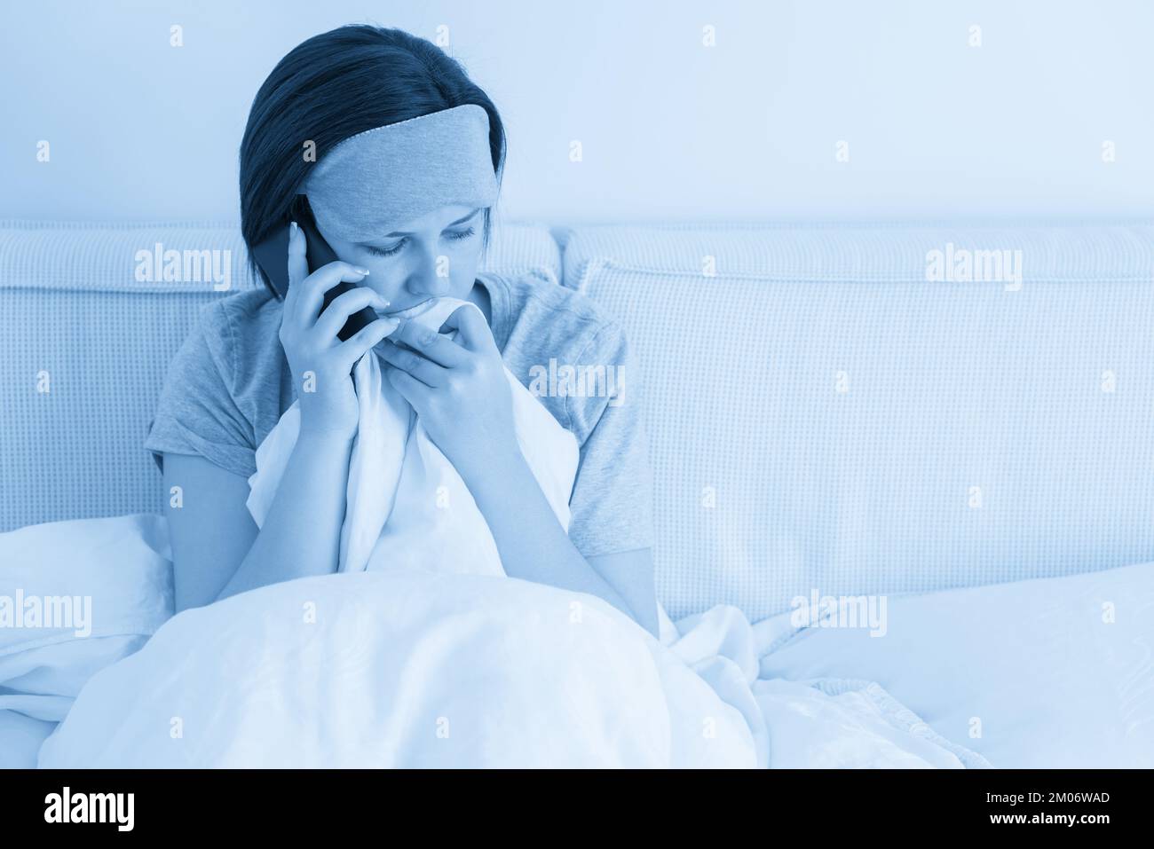 Donna triste che indossa la maschera del sonno con lo smartphone mentre si  siede nel letto coperto di piumino Foto stock - Alamy
