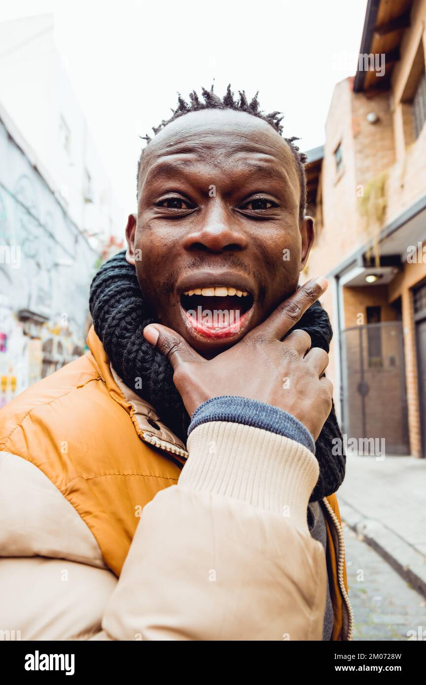 felice giovane uomo africano che fa bella espressione sorridendo guardando  la macchina fotografica, indossa abiti invernali, sciarpa nera e giacca color  senape Foto stock - Alamy