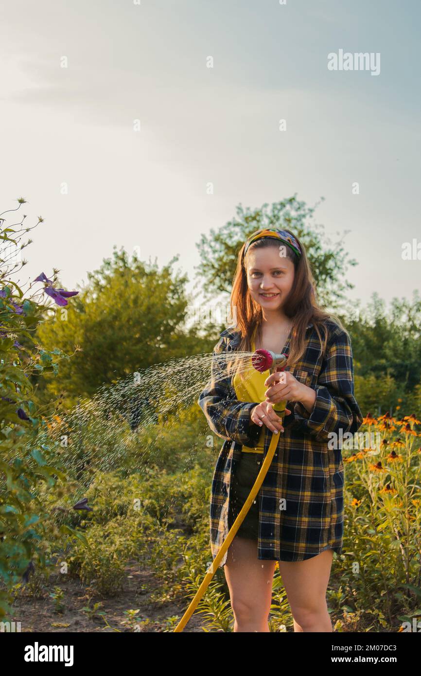 giovane ragazza graziosa che si diverte nel giardino innaffiare le piante con un tubo. Sorridendo mentre prende un hobby favorito Foto Stock