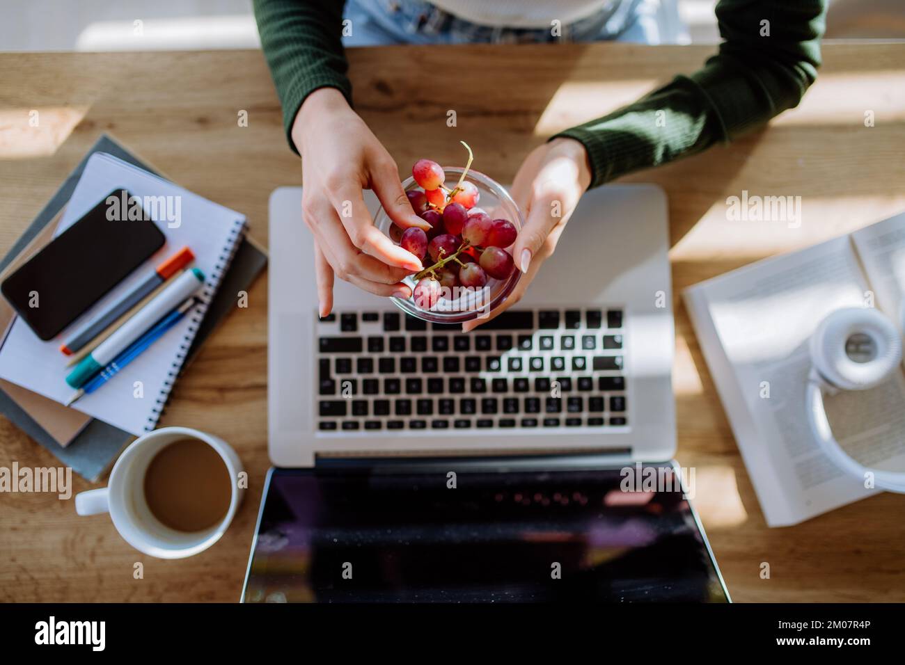 Vista dall'alto della donna che tiene il recipiente con uva sopra la scrivania con computer, diario e smartphone. Concetto di equilibrio tra vita lavorativa e vita privata. Foto Stock