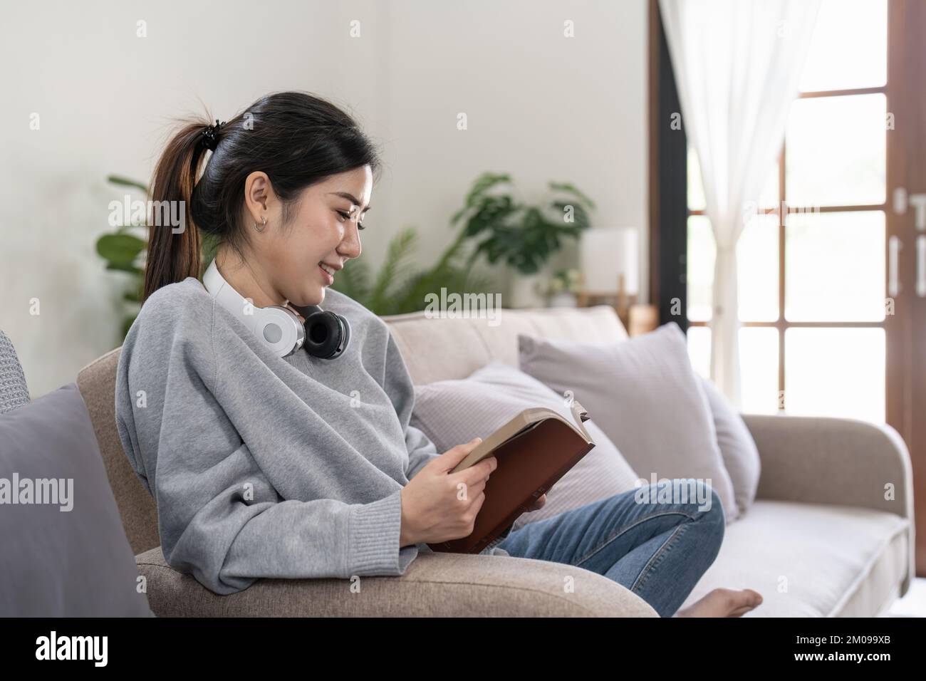 Giovane bella studentessa asiatica seduta sul divano lettura libro a casa. Foto Stock