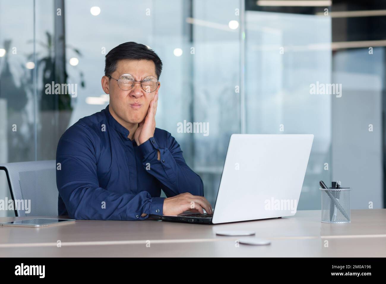 Asiatico malato in ufficio, uomo d'affari ha un mal di denti, uomo seduto in ufficio all'interno di un edificio che lavora al lavoro utilizzando il laptop, rovesciato capo in camicia vicino alla finestra. Foto Stock