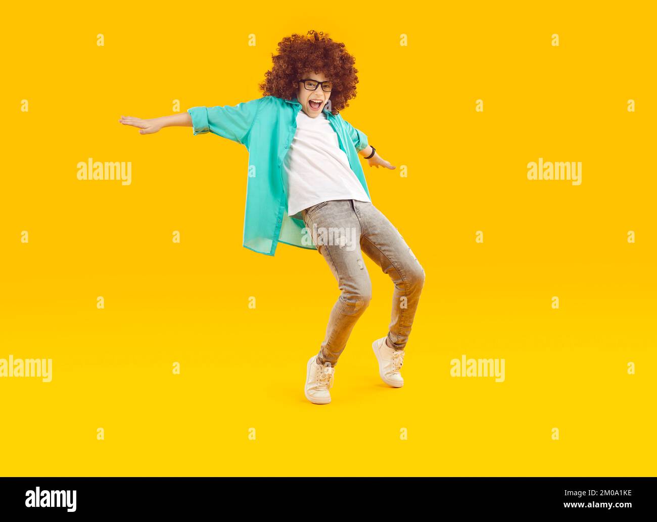 Felice bambino divertente che indossa una parrucca curly ballare su uno sfondo giallo arancione studio Foto Stock