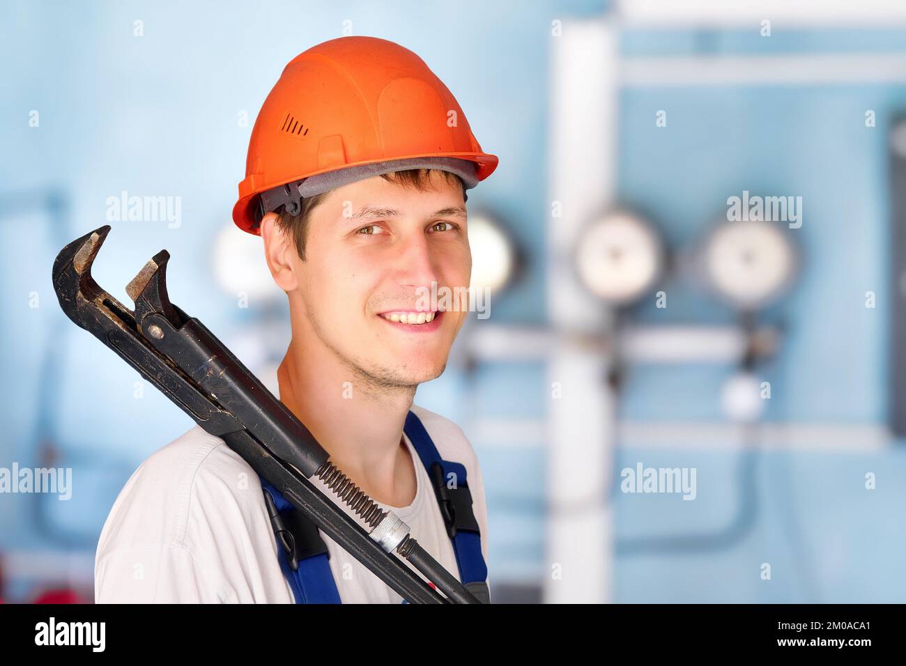 L'idraulico industriale positivo o l'operatore nel casco tiene la chiave regolabile grande sulla sua spalla e sorride. Ritratto in interni sullo sfondo di sensori e manometri in sfocatura. Foto Stock