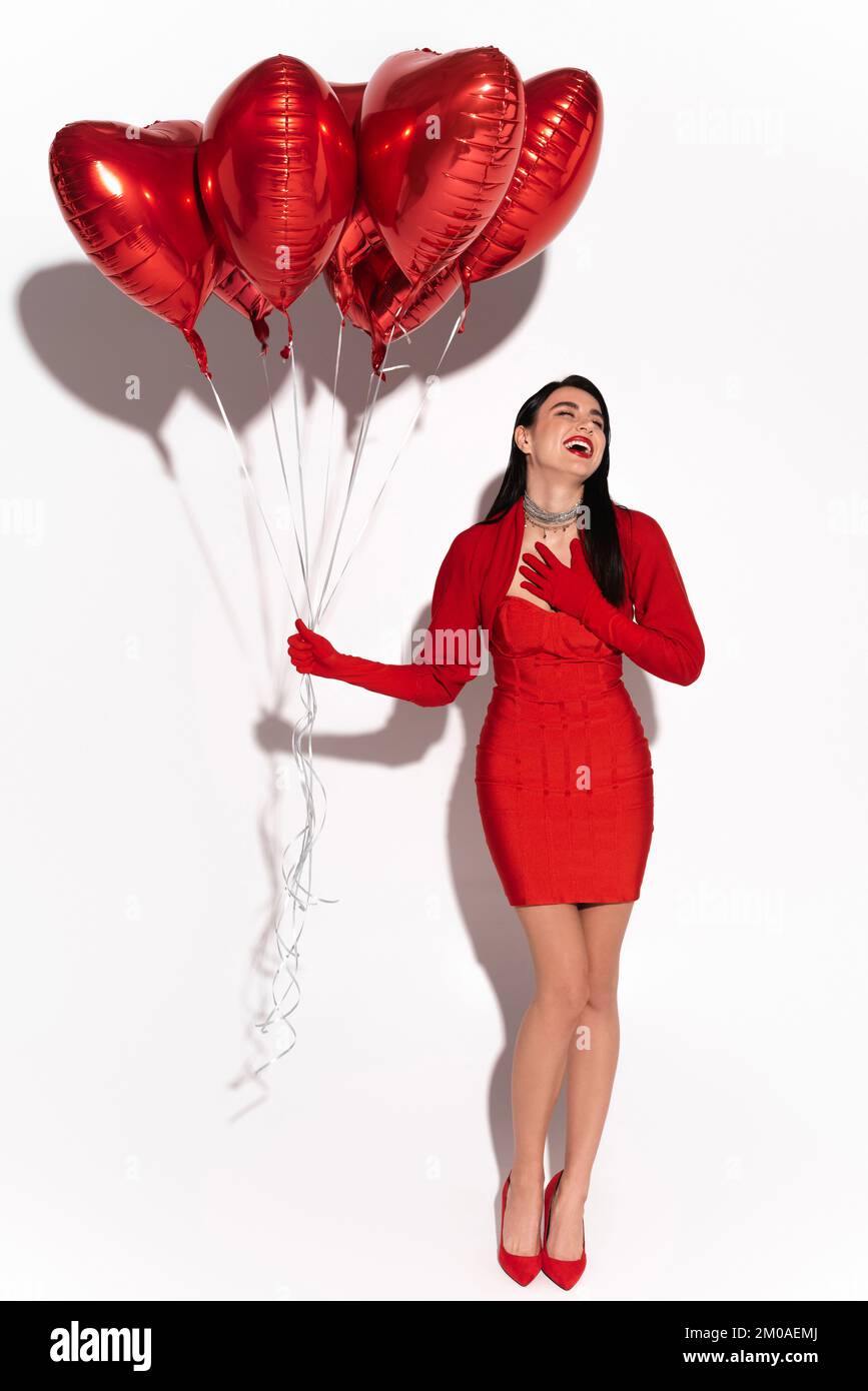 Donna elegante in abiti rossi ridendo e tenendo palloncini a forma di cuore su sfondo bianco con ombra Foto Stock