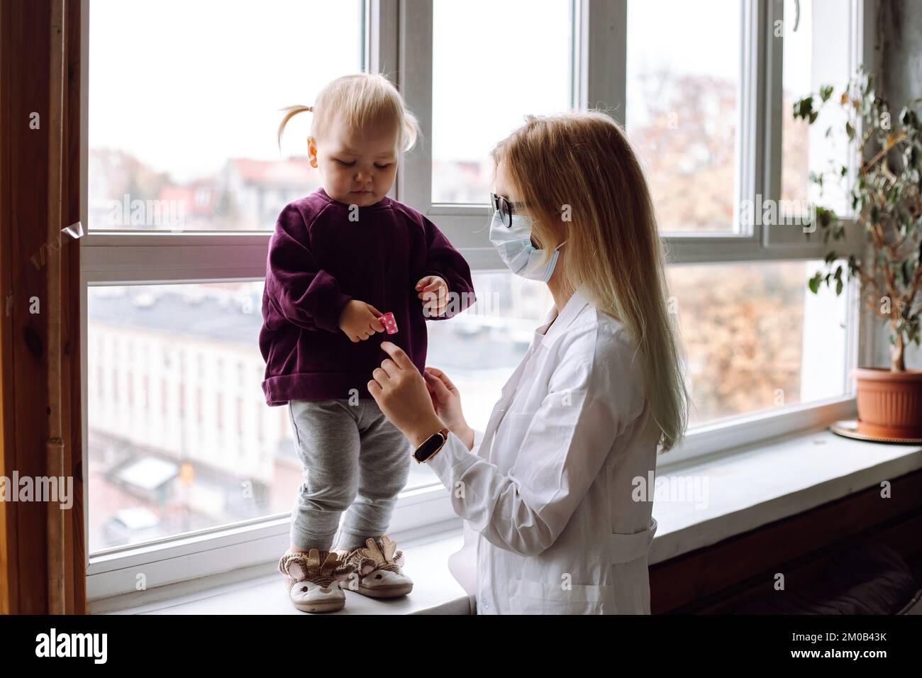 Piccola ragazza bionda in piedi sul windowsill, guardando il giocattolo in mano vicino al medico dei bambini che indossa uniforme bianca a casa. Foto Stock