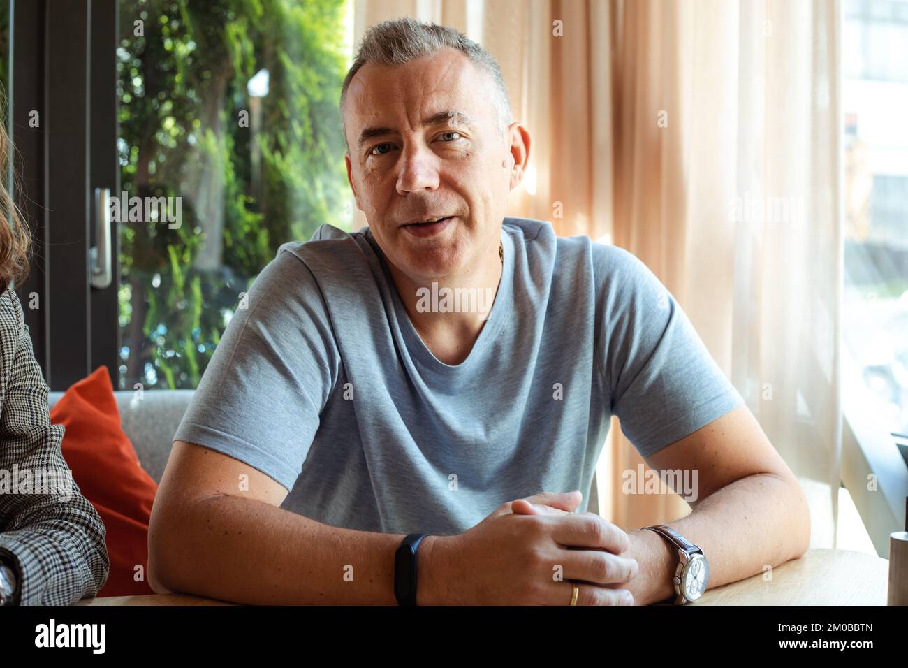 Sorridente uomo grizzled sicuro in t-shirt grigia, con smartwatch, guardare la macchina fotografica, sedersi al tavolo vicino alla finestra. Intervista lavoro Foto Stock