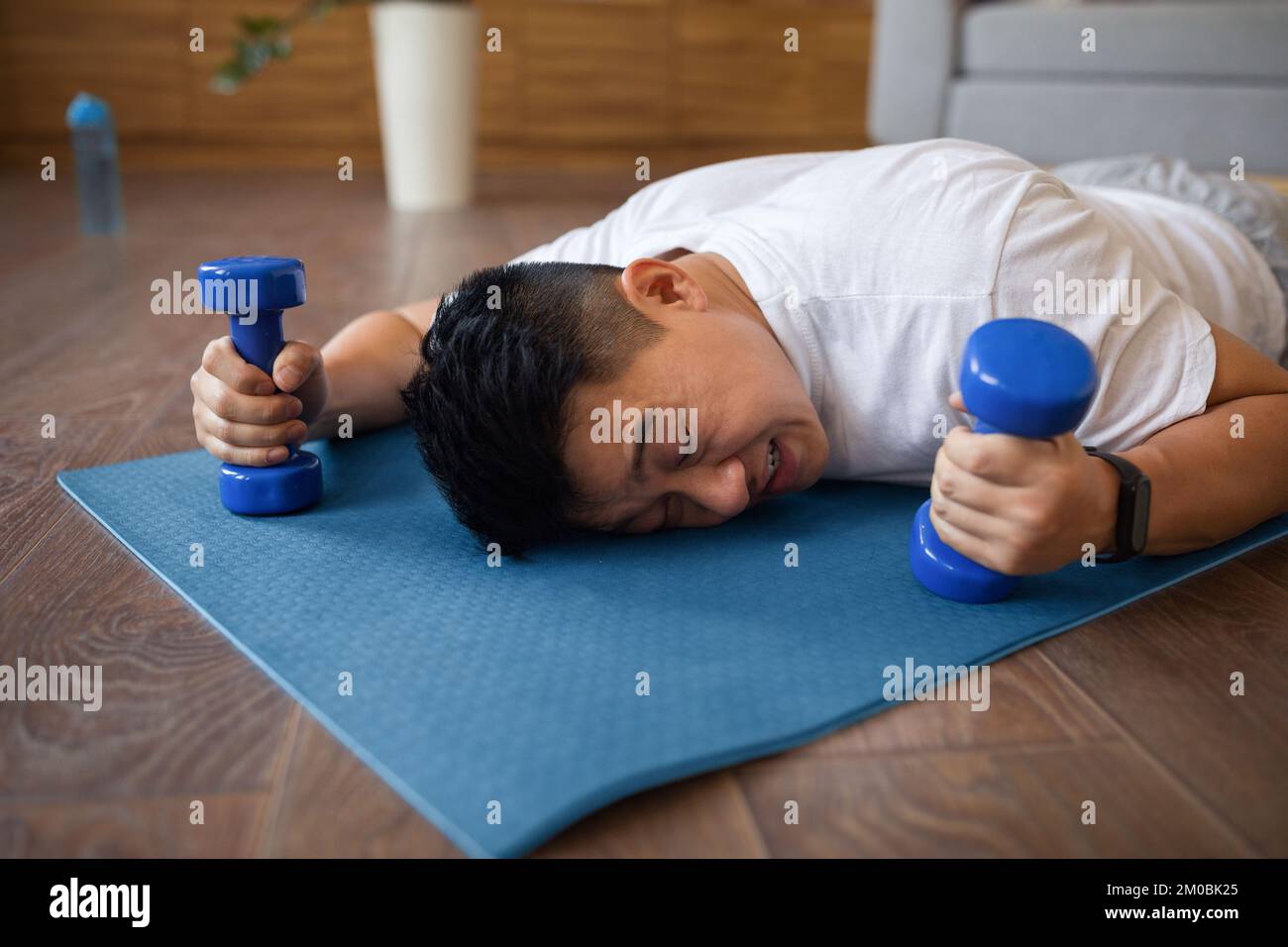 Uomo maturo asiatico stanco sdraiato sul tappetino fitness con manubri in mano, riposante dopo l'allenamento domestico nel soggiorno Foto Stock