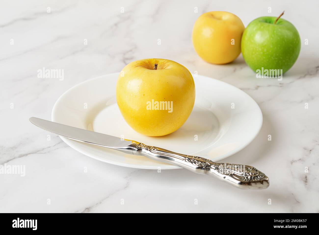 Mela gialla matura e coltello su un piatto bianco su fondo di marmo. Frutta fresca e succosa per un'alimentazione sana delle vitamine. Snack vegetariano antiossidante. Foto Stock