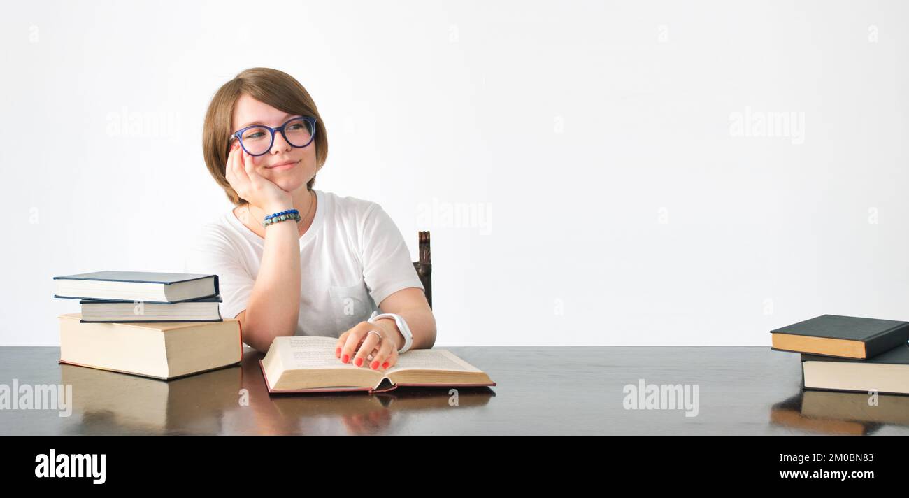 Una giovane studentessa intelligente ispirata seduta e studio con libri su una scrivania, sognando l'ispirazione e isolato contro un muro bianco ba Foto Stock