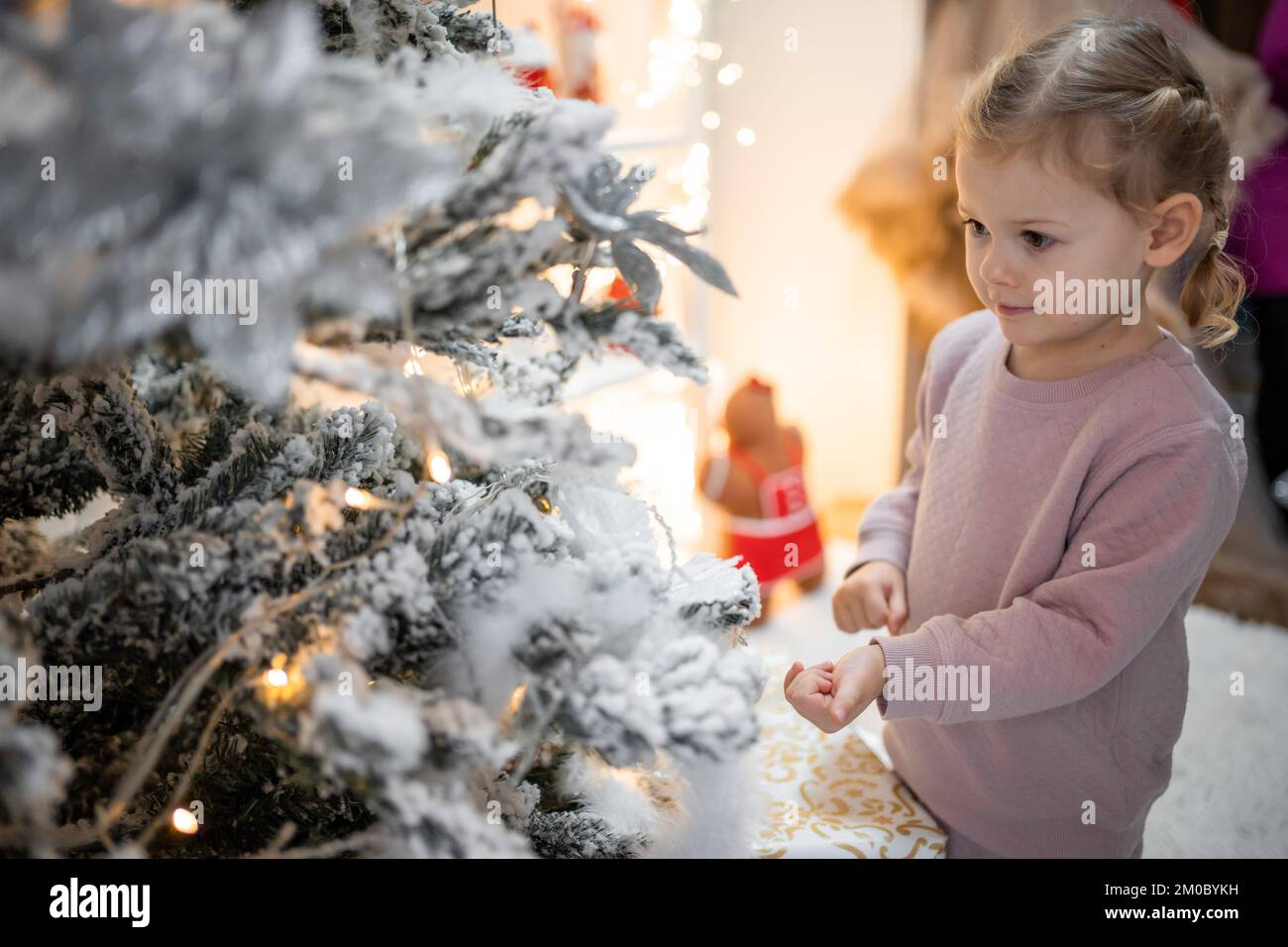 Molto bella piccola ragazza bionda affascinante in pigiama vicino alberi di Natale in luminoso interno della casa Foto Stock