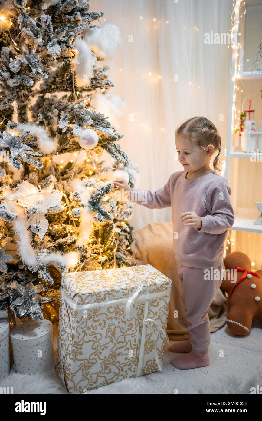 Molto bella piccola ragazza bionda affascinante in pigiama vicino alberi di Natale in luminoso interno della casa Foto Stock