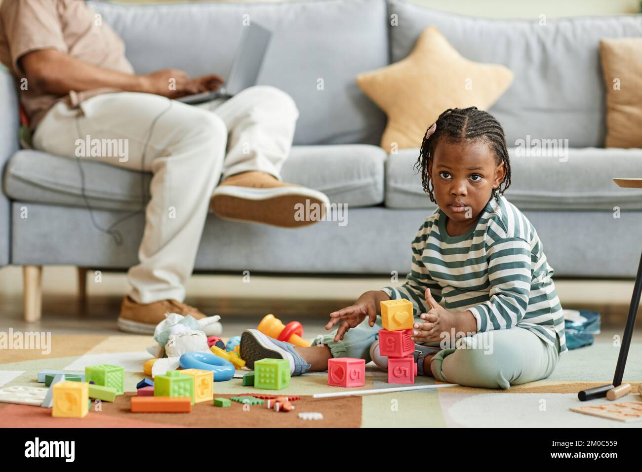 Ritratto completo di carino bambino nero giocare con blocchi giocattolo mentre si siede sul pavimento a casa con padre che lavora in background, copia spazio Foto Stock