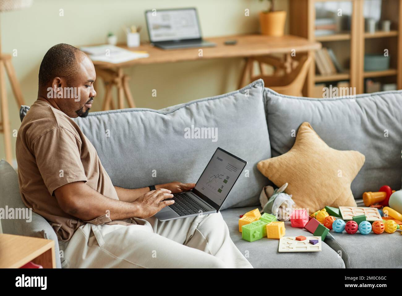Vista laterale ritratto di un singolo padre che lavora da casa e che utilizza il computer portatile sul divano con giocattoli, spazio copia Foto Stock