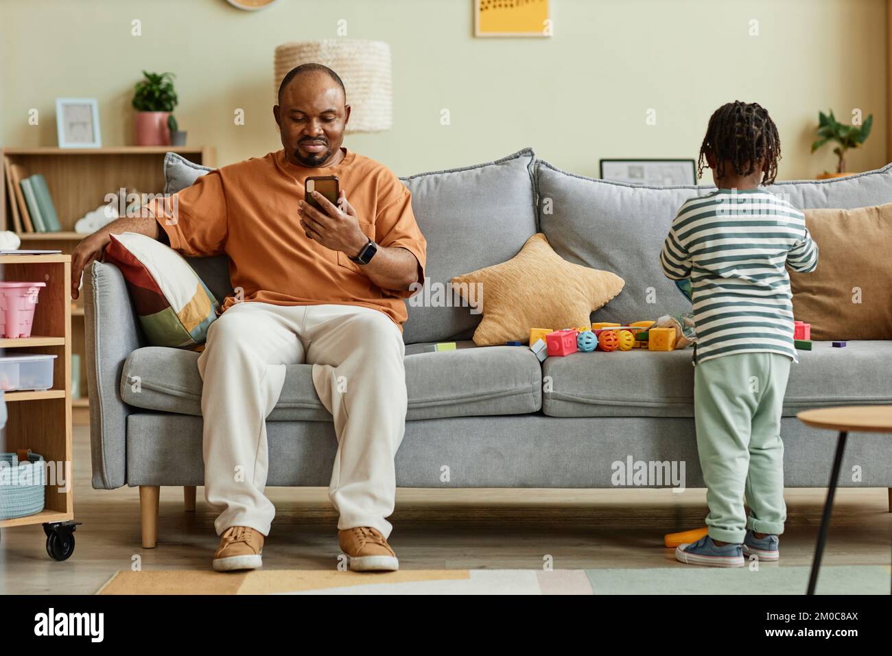 Ritratto completo del padre che usa lo smartphone con il figlio che gioca sul divano a casa, spazio copia Foto Stock