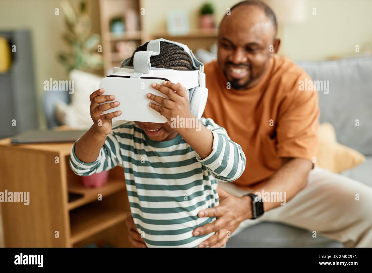 Ritratto di un simpatico bambino nero che indossa un visore VR e che si diverte con la realtà virtuale per i bambini con padre sullo sfondo Foto Stock