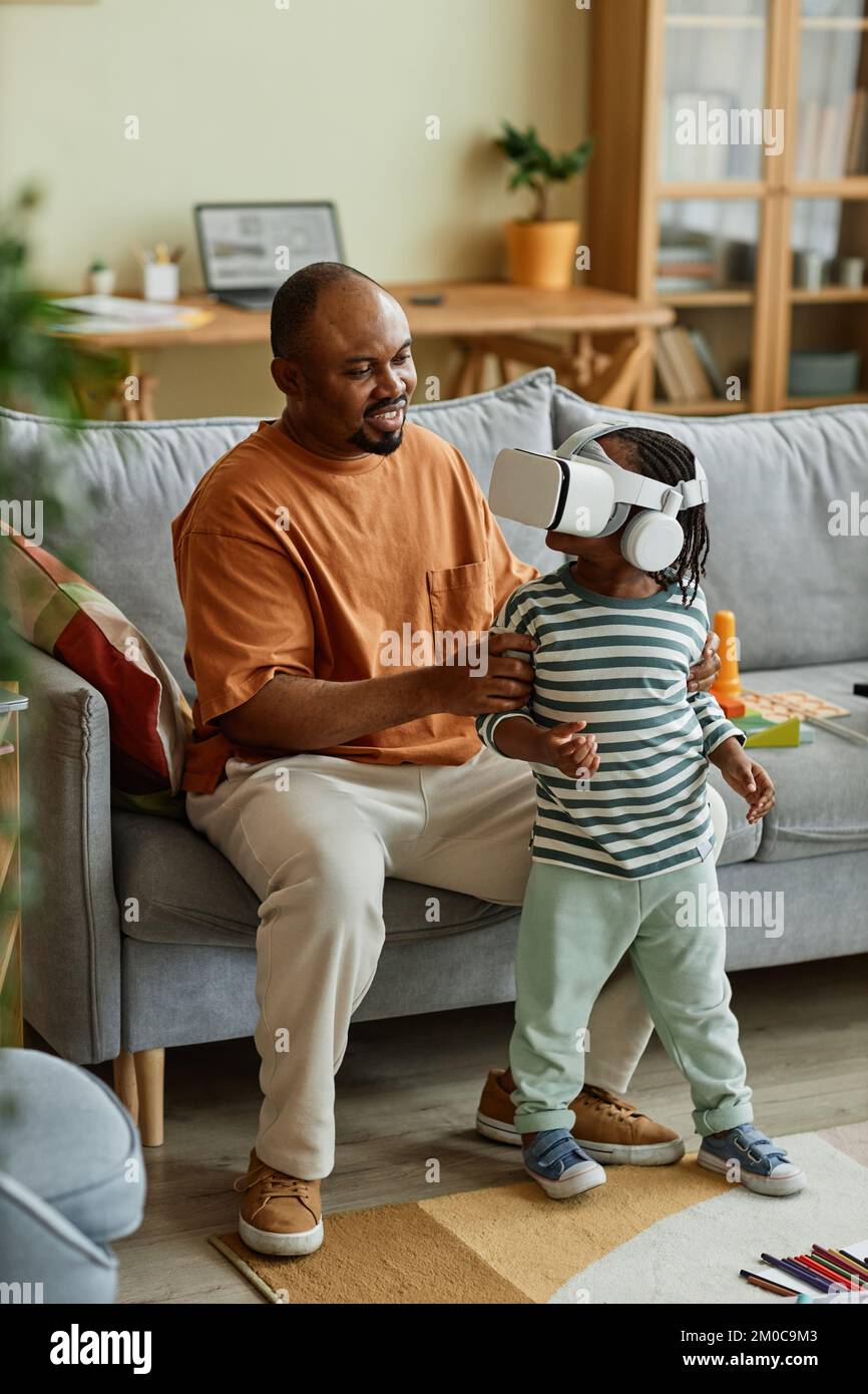 Ritratto verticale di un simpatico bambino nero che indossa un visore VR e ti offre l'intrattenimento in realtà virtuale con il supporto del padre Foto Stock