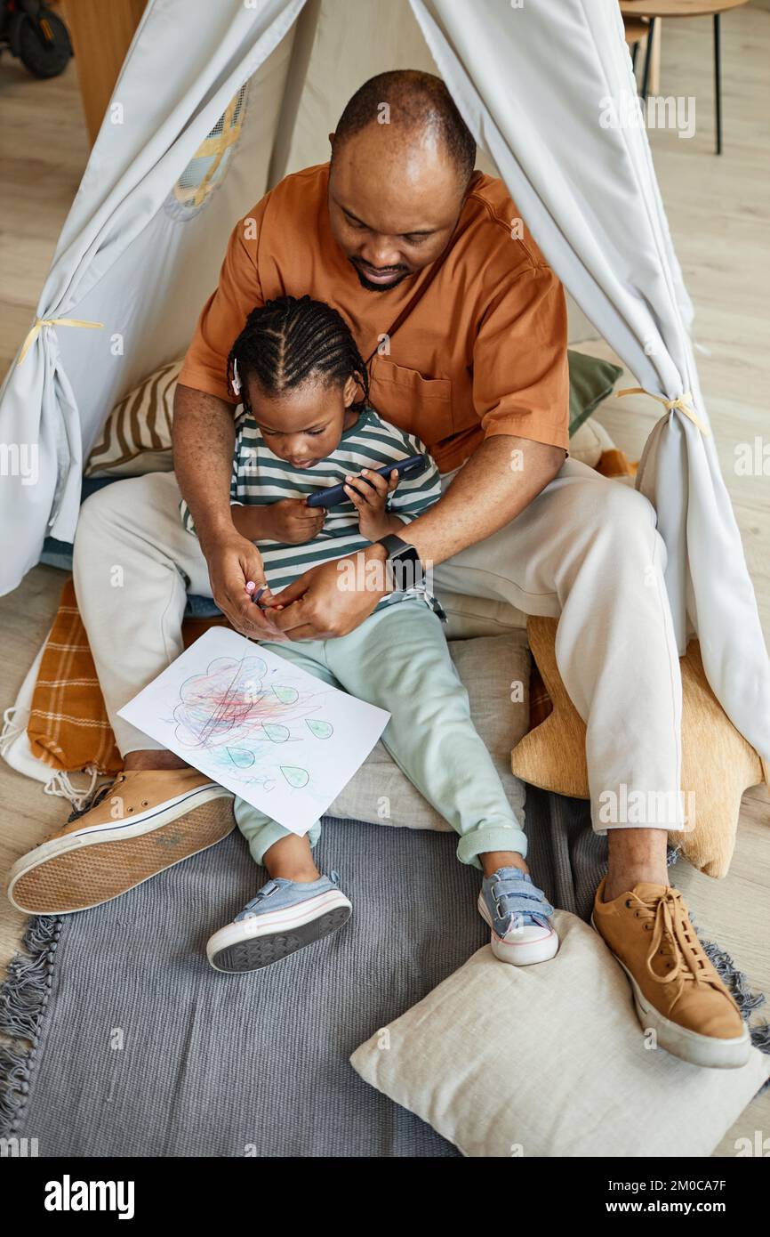 Verticale ritratto a tutta lunghezza di amorevole padre nero con bambino che si disegna insieme in tenda da gioco Foto Stock