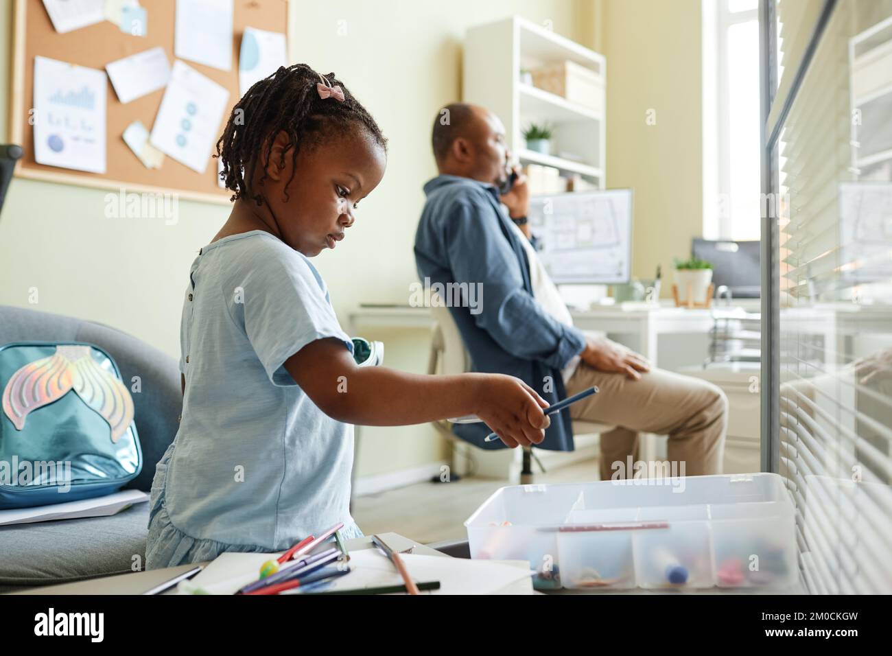Vista laterale ritratto di bambina nera scelta matite colorate mentre disegno in ufficio con padre che lavora in background Foto Stock