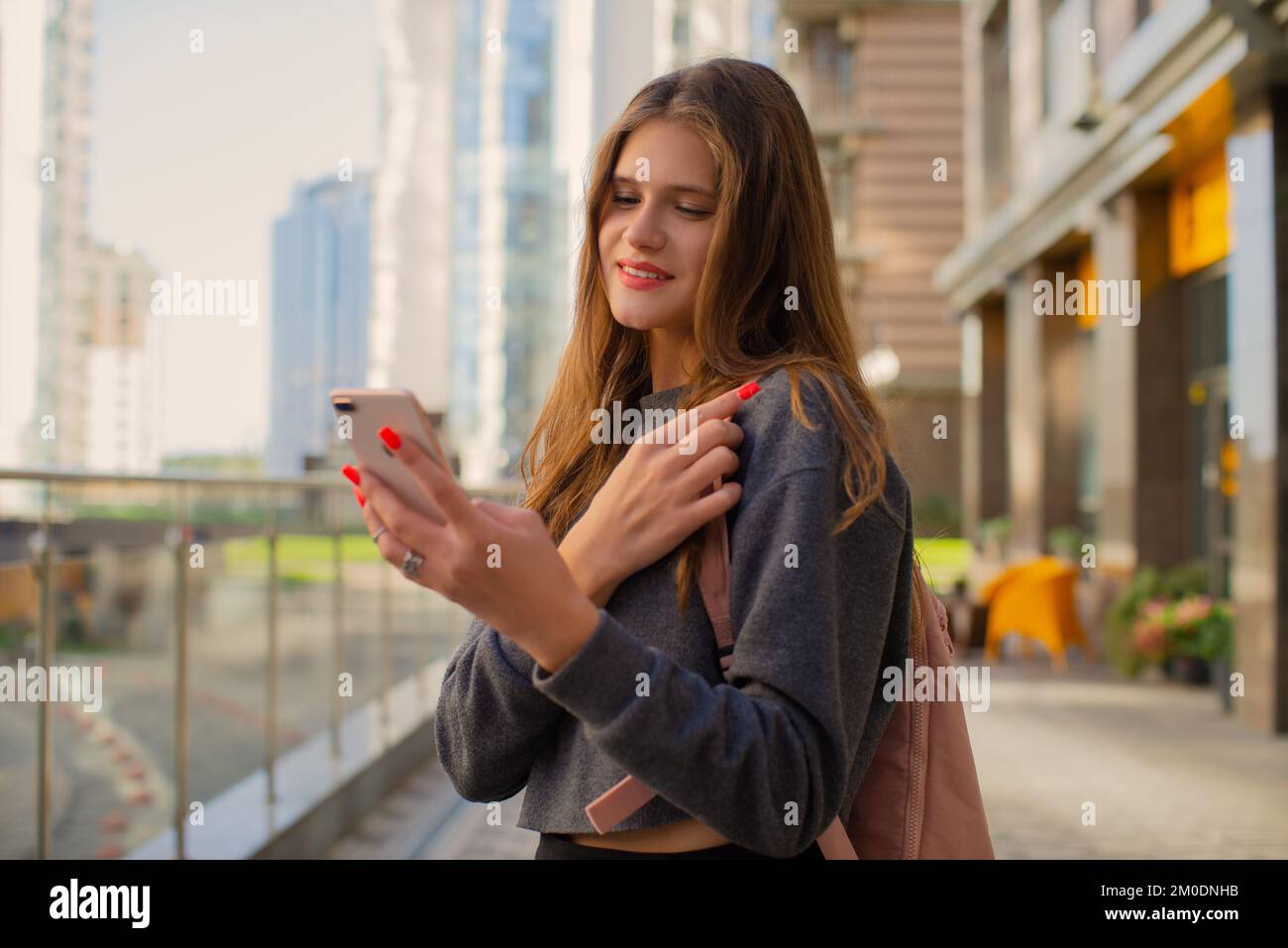 Donna casual sorridente con zaino che utilizza il telefono cellulare iPhone in piedi per strada in centro all'aperto Foto Stock