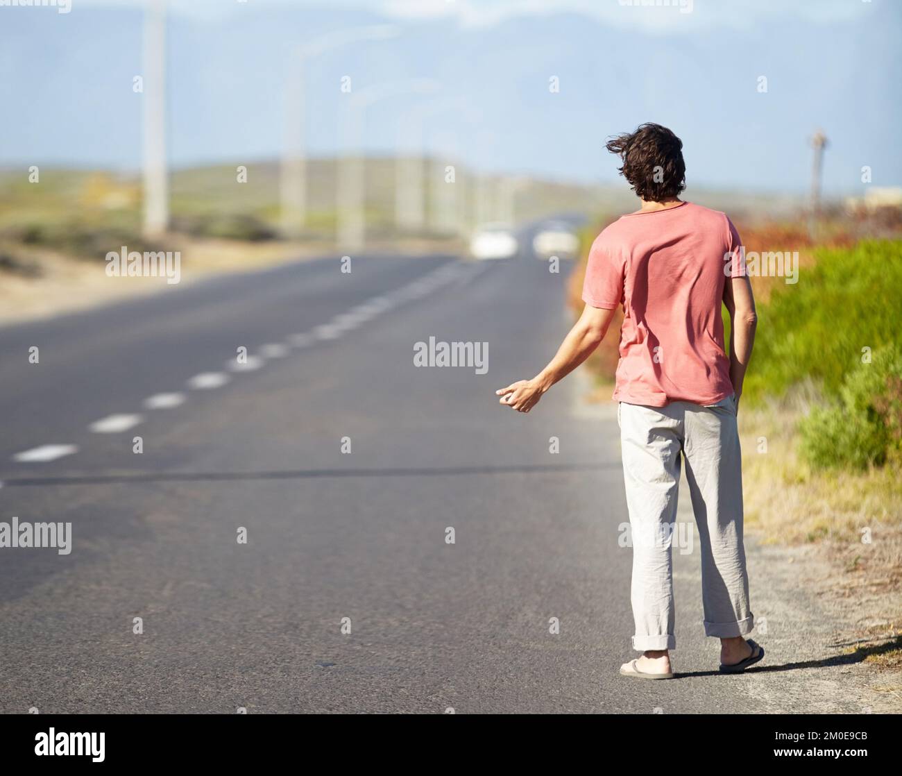 Indovino che Ill debba hitchhike. Un giovane che cerca di agganciare un giro mentre cammina lungo una strada deserta. Foto Stock