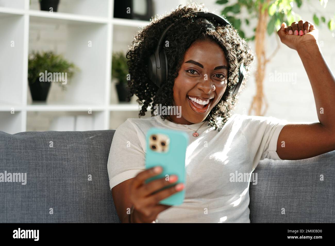 Donna africana sorridente seduta sul divano a casa e con lo smartphone Foto Stock