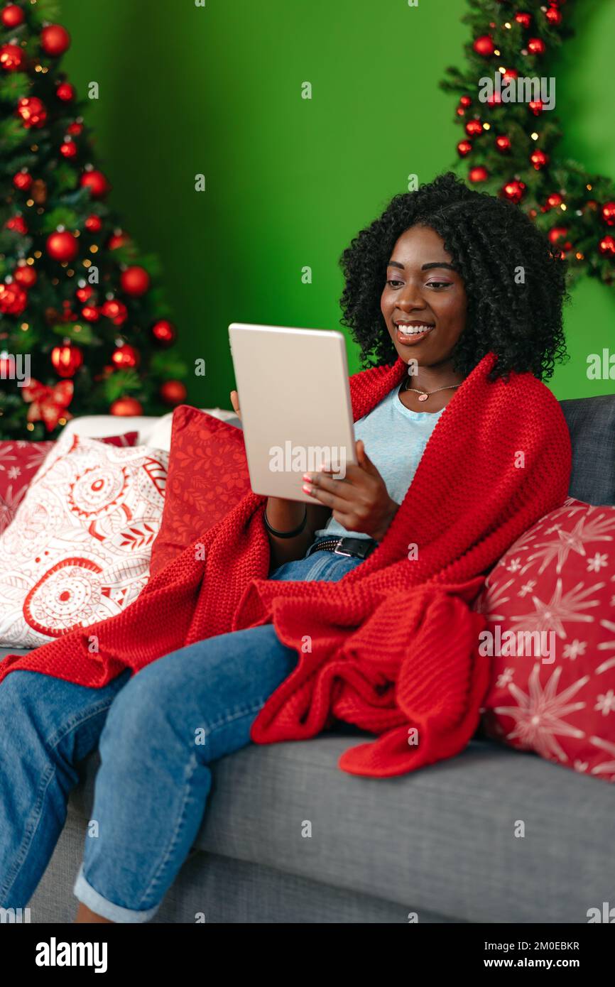 Giovane donna africana che usa un tablet digitale e sorridente mentre riposa a casa decorata per Natale. Foto Stock