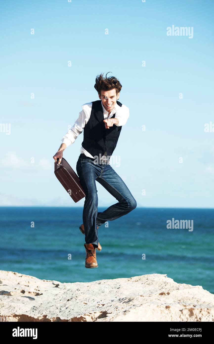 Anche voi potreste avere questa libertà. Bel giovane uomo che tiene una valigetta saltando in aria con l'oceano sullo sfondo. Foto Stock