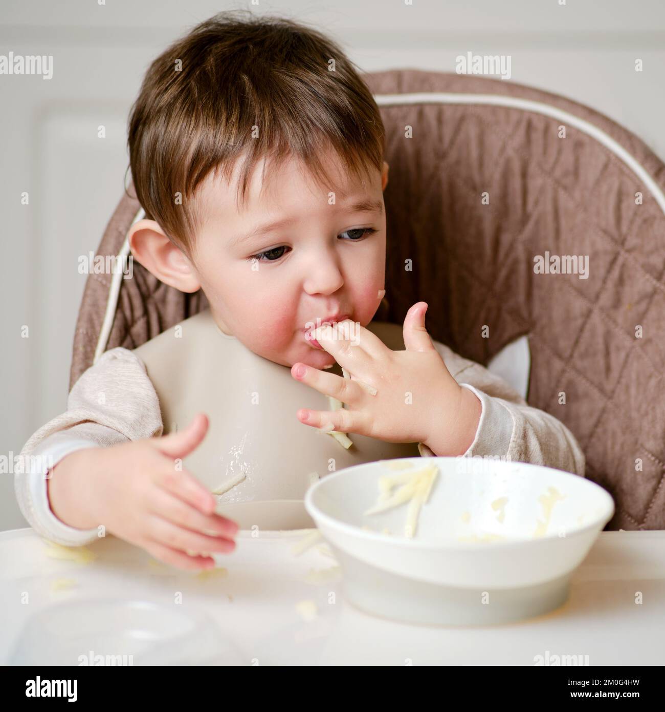 Un bambino divertente sta mangiando una mela grattugiata con la bocca piena  mentre si siede su una sedia da cucina. Il bambino affamato infila il cibo  in bocca, umorismo. Bambino di età