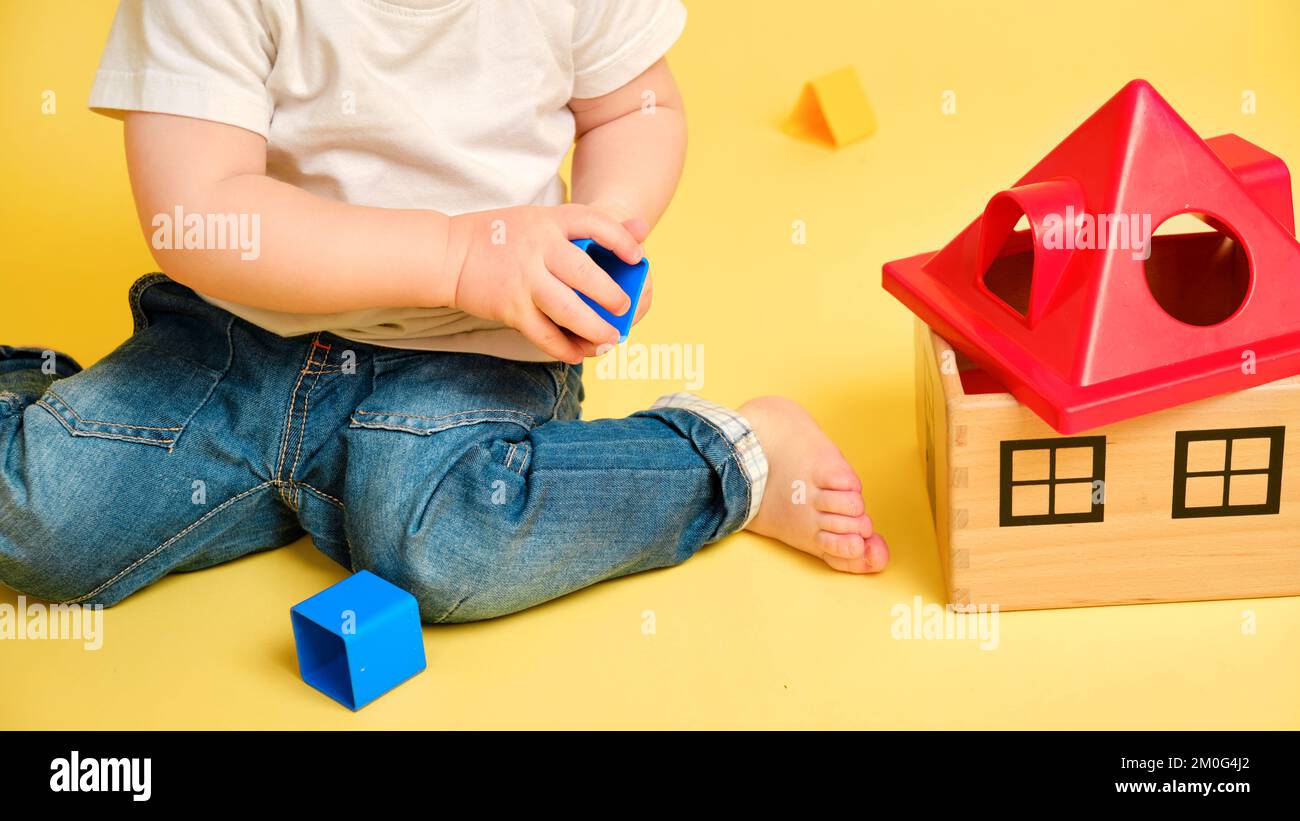 Il bambino del toddler sta giocando i giochi educativi di logica del selezionatore con le forme geometriche su uno sfondo giallo dello studio. Felice gioco del bambino con hous del giocattolo educativo Foto Stock