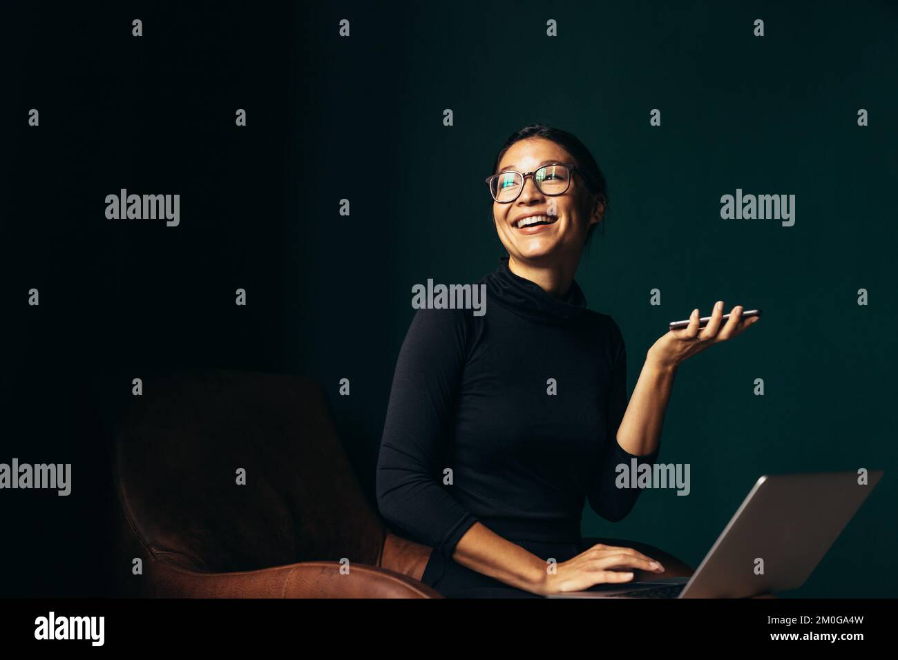 Giovane donna con notebook che parla con vivavoce e sorridente. Donna asiatica seduta su sedia con cellulare e guardando lontano. Foto Stock