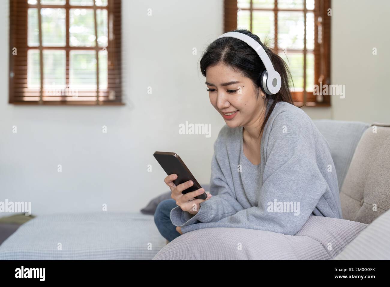 Donna asiatica ascoltare musica con telefono cellulare e cuffie. Calma casa fine settimana, rilassarsi mentre si siede sul divano, godersi la musica preferita, musica popolare, utilizzare moderno Foto Stock