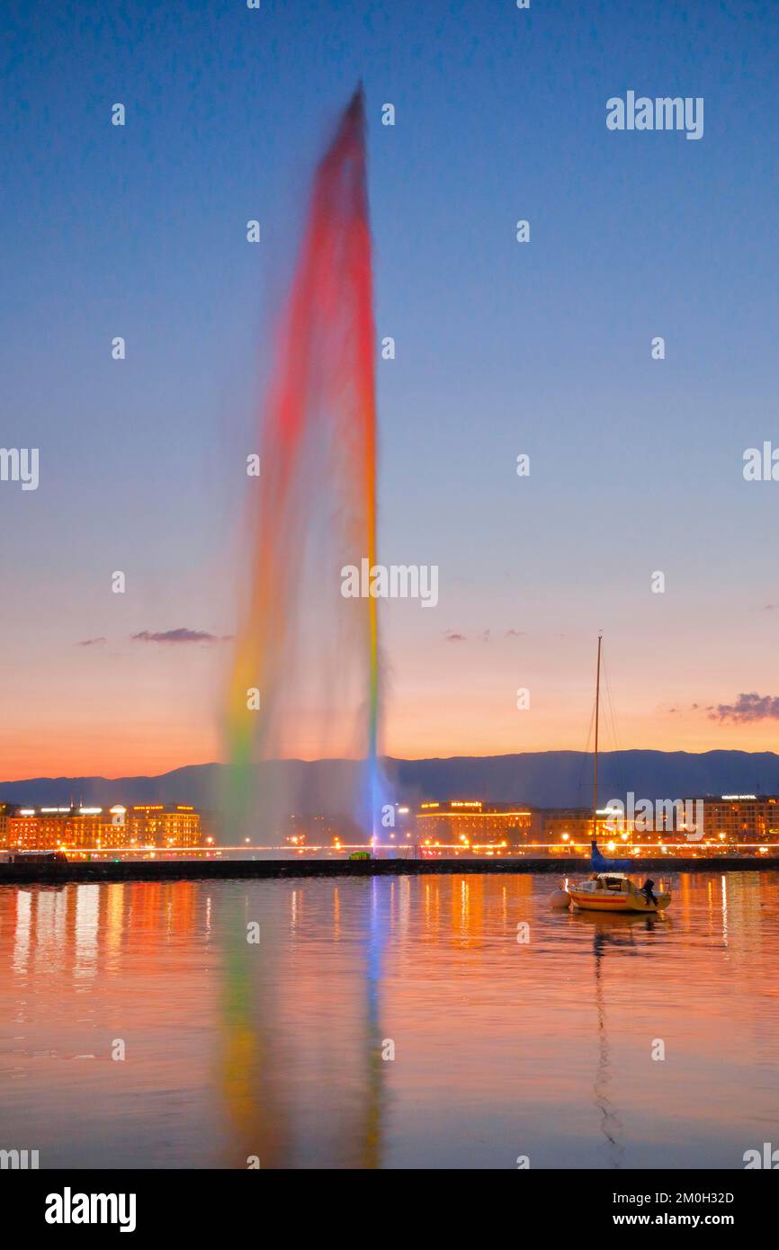 Il Jet d'eau illuminato al crepuscolo è il punto di riferimento nel bacino del Lago di Ginevra, con barche a vela in primo piano e la passeggiata sul lago illuminata a Nig Foto Stock