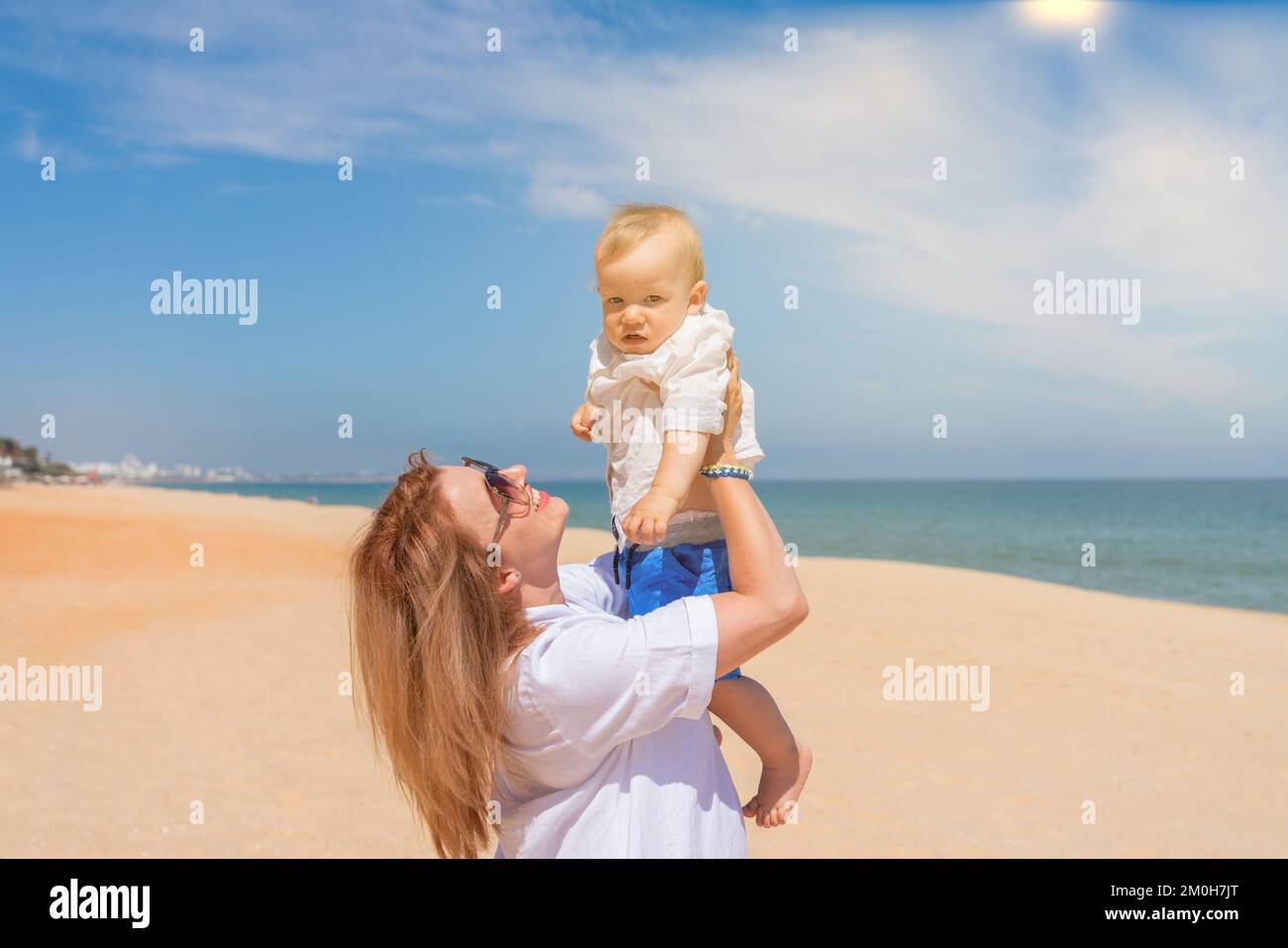 Giovane madre in un vestito bianco sulla spiaggia vicino al mare tiene il suo figlio bambino nelle braccia e gioisce al sole. Concetto di maternità e amore per i bambini. Foto Stock