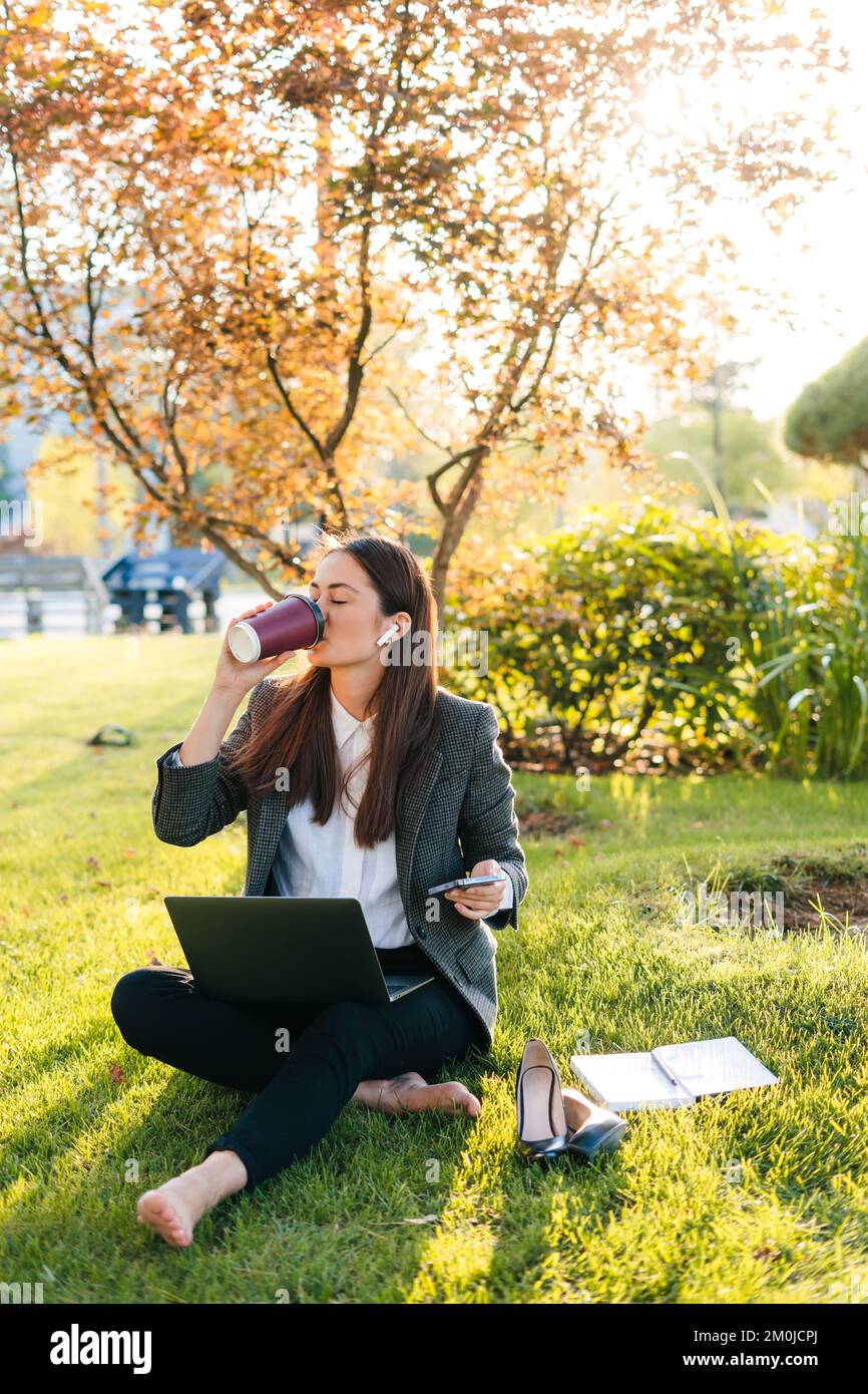 Bella donna che si rilassa bevendo una tazza di caffè dopo il lavoro duro e utilizzando il computer portatile mentre si siede sull'erba verde nel parco. Tecnologia e libertà Foto Stock
